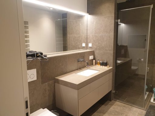 Modernes Badezimmer mit maßgefertigtem Waschtisch mit weißen Fronten, großflächigem Spiegel und integrierter Beleuchtung.